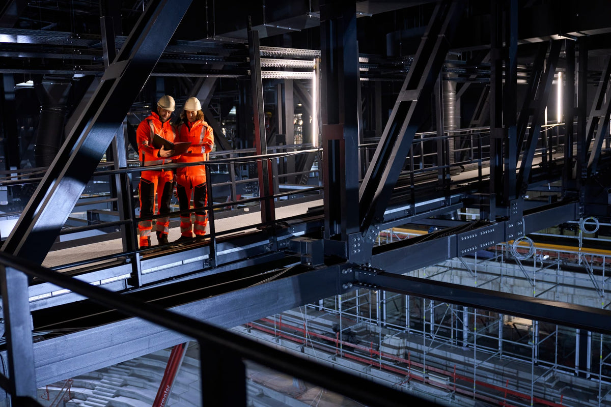 Zwei Mitarbeitende in orangefarbener MASCOT-Warnschutzkleidung auf einem Steg in einer Industriehalle